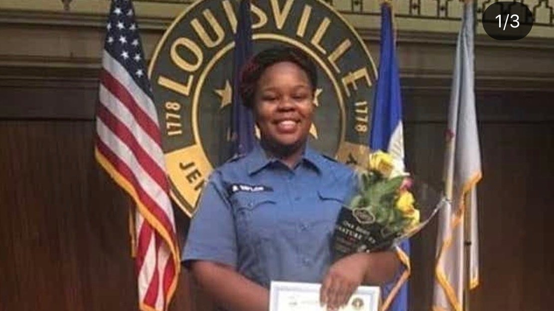 Breonna Taylor stands with a bouquet of flowers in a blue uniform in front of a U.S. flag and seal of the city of Louisville, Ketucky.